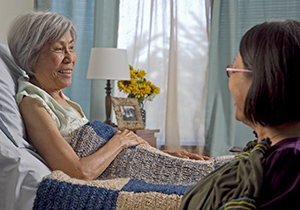 Woman lying in bed, woman sitting next to bed.